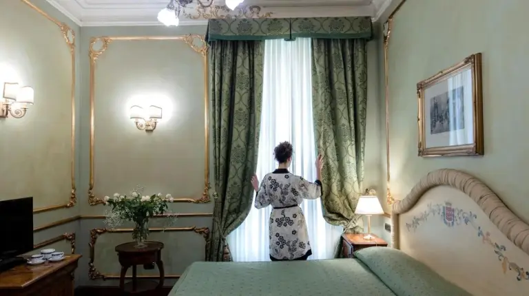 eine Frau steht in einem Schlafzimmer und blickt aus dem Fenster im Grand Hotel Wagner in Palermo