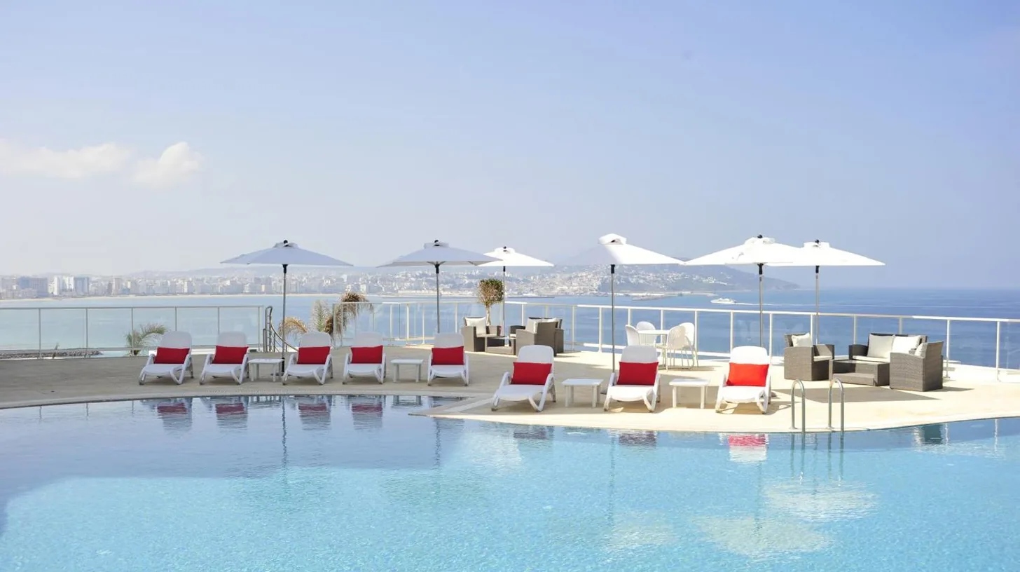 a swimming pool with chairs and tables and the ocean at Hotel Farah Tanger in Tangier