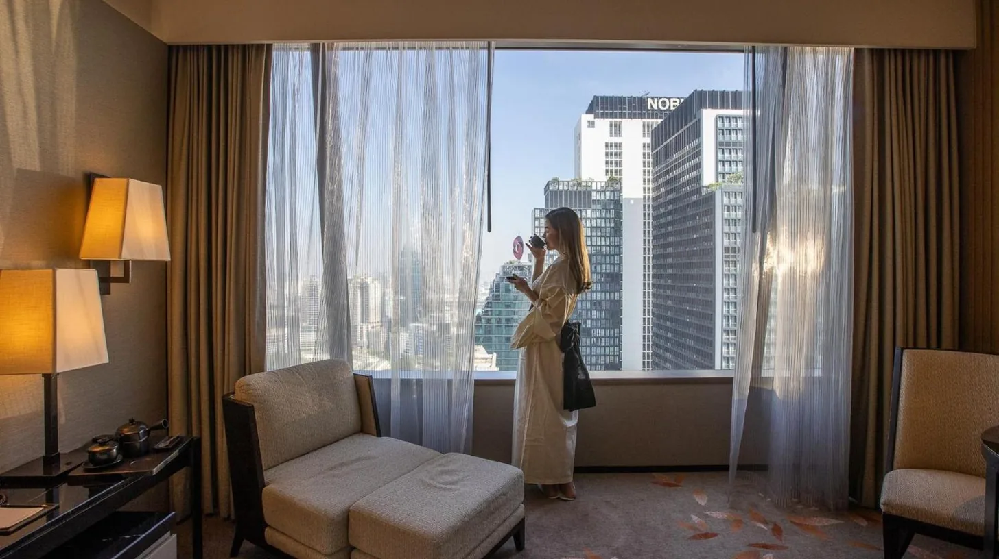 a woman standing in a hotel room looking out the window at The Okura Prestige Bangkok in Bangkok